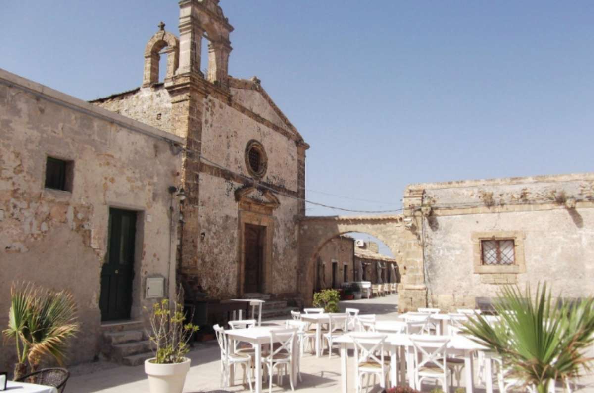 A sunlit courtyard in Sicily with white outdoor tables, surrounded by weathered stone buildings and a rustic church, Italy