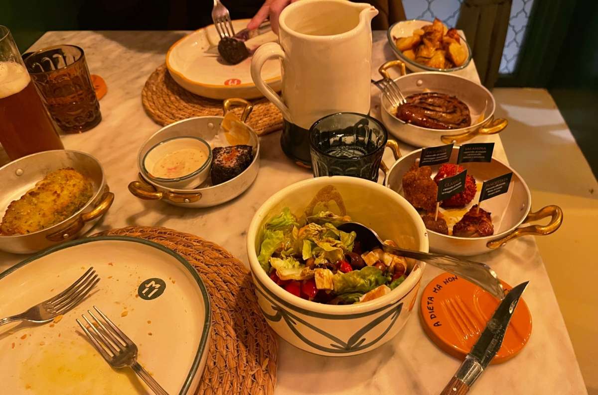 A table at a restaurant in Modena, Italy, filled with various dishes including salad, grilled sausage, croquettes, dipping sauce, and dessert, photo by Next Level of Travel