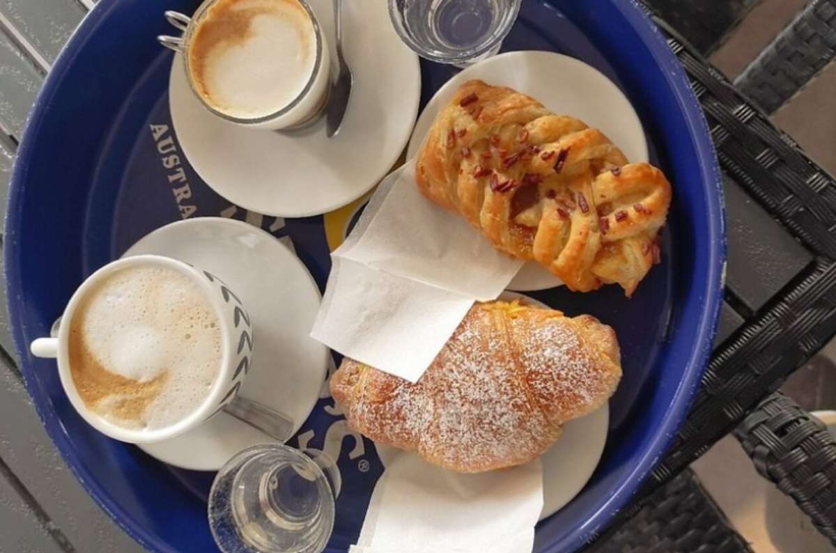 A tray with two cappuccinos, two glasses of water, a croissant dusted with powdered sugar, and a braided pastry, Italian breakfast 