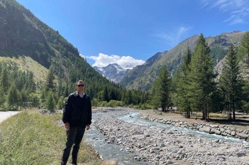 A tourist standing near the river in Gran Paradiso National Park, mountains and forests visible in the background, Italy, photo by Next Level of Travel