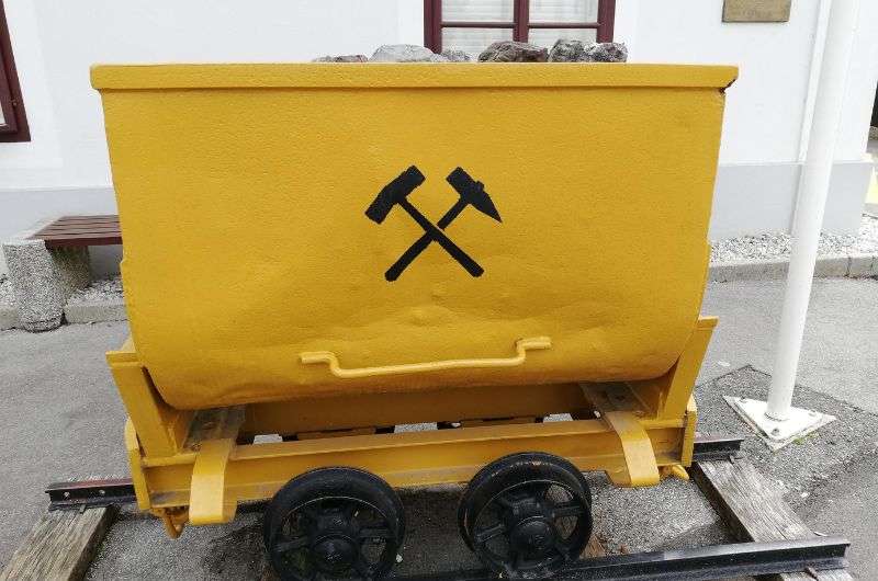 A yellow mining cart filled with rocks, featuring a black hammer and pickaxe symbol, standing on tracks outside Schwaz Silver Mine, Austria