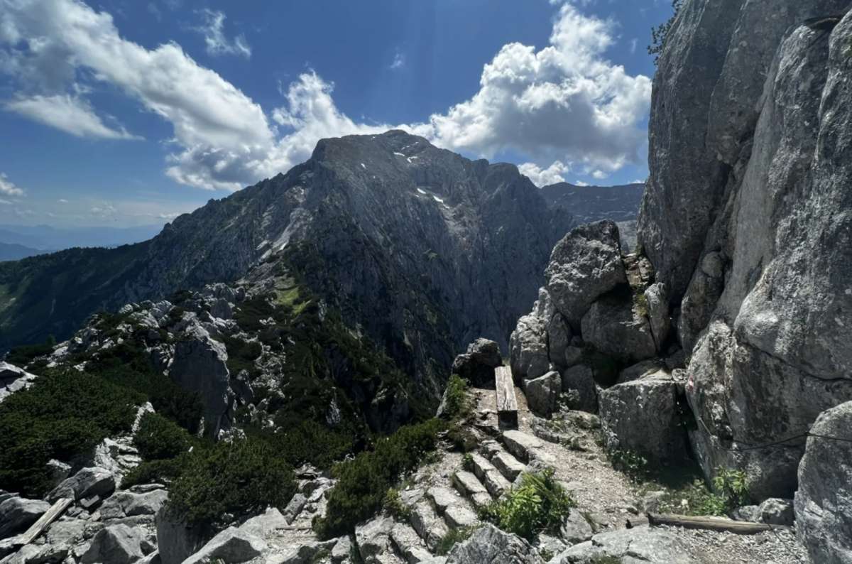 Rocky mountain trail leading to a wooden bench with a breathtaking view of rugged peaks and dramatic cliffs, under a partly cloudy blue sky near Eagle's Nest (Kehlsteinhaus), Germany