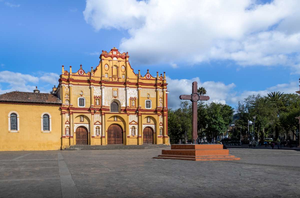 a photo of the yellow baroque facade of the Cathedral of San Cristóbal de las Casas with a stone cross in the main plaza, San Cristóbal de las Casas, Mexico, photo by Next Level of Travel