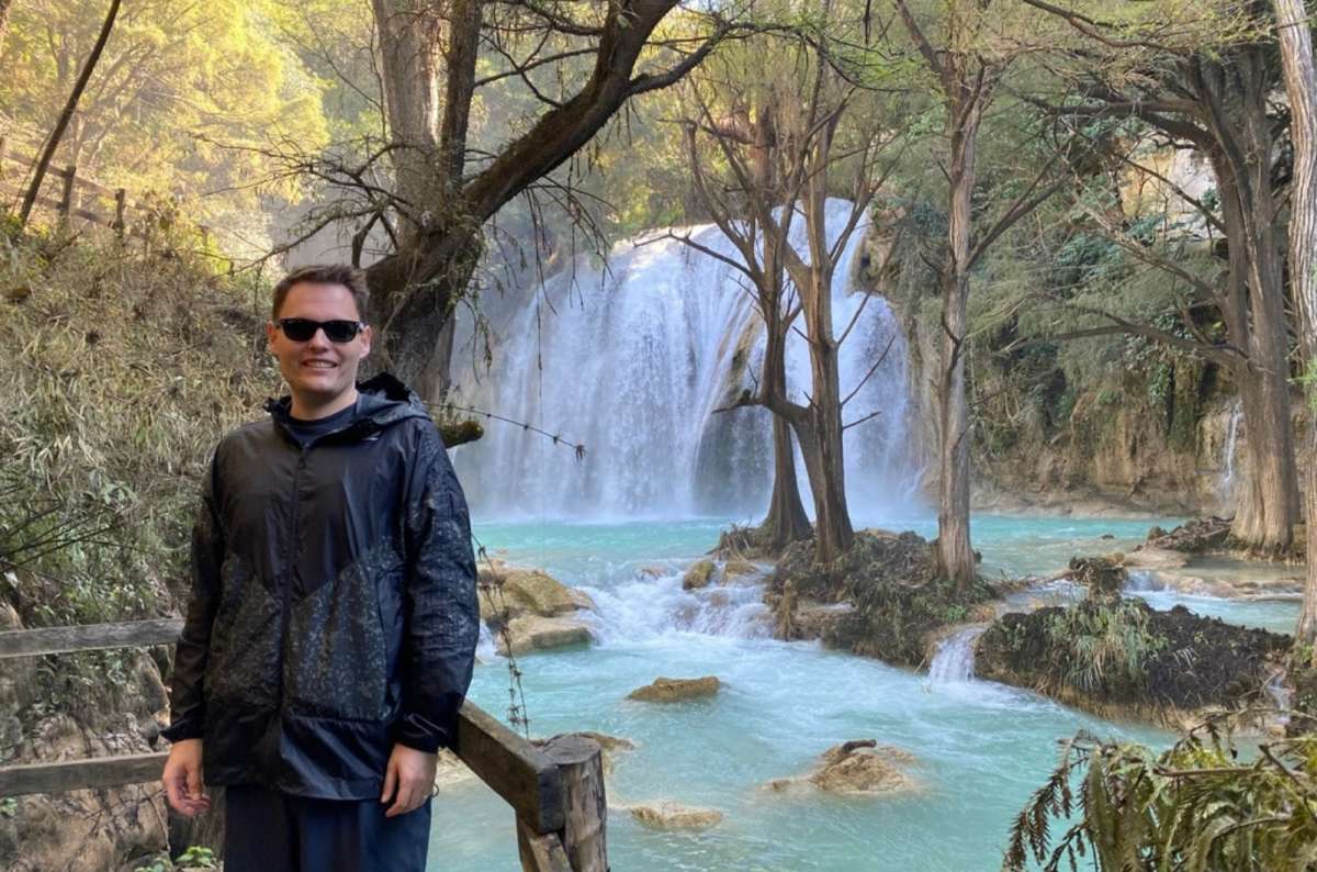 a photo of a tourist posing in front of the El Chiflon waterfall in Chiapas, with green trees and the waterfall itself in the background, Mexico, photo by Next Level of Travel