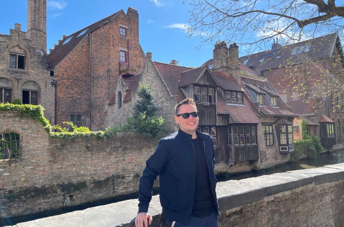 Smiling man posing by the canal with medieval houses in Bruges’ historic center, a must-see spot when searching for what to see in Bruges, photo by Next Level of Travel