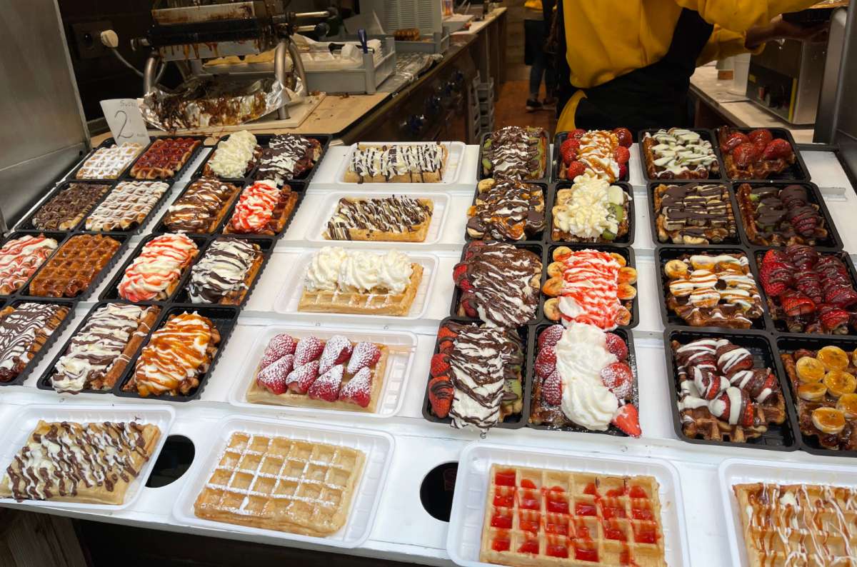 A variety of Belgian waffles topped with whipped cream, chocolate, fruit, and sauces on display at a street food stall in Belgium, photo by Next Level of Travel