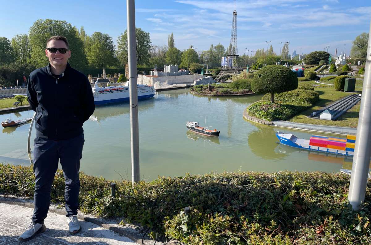 a photo of a tourist posing in front of Mini-Europe on a sunny spring day with a miniature of the Eiffel tower visible in the background, Brussels, Belgium, photo by Next Level of Travel