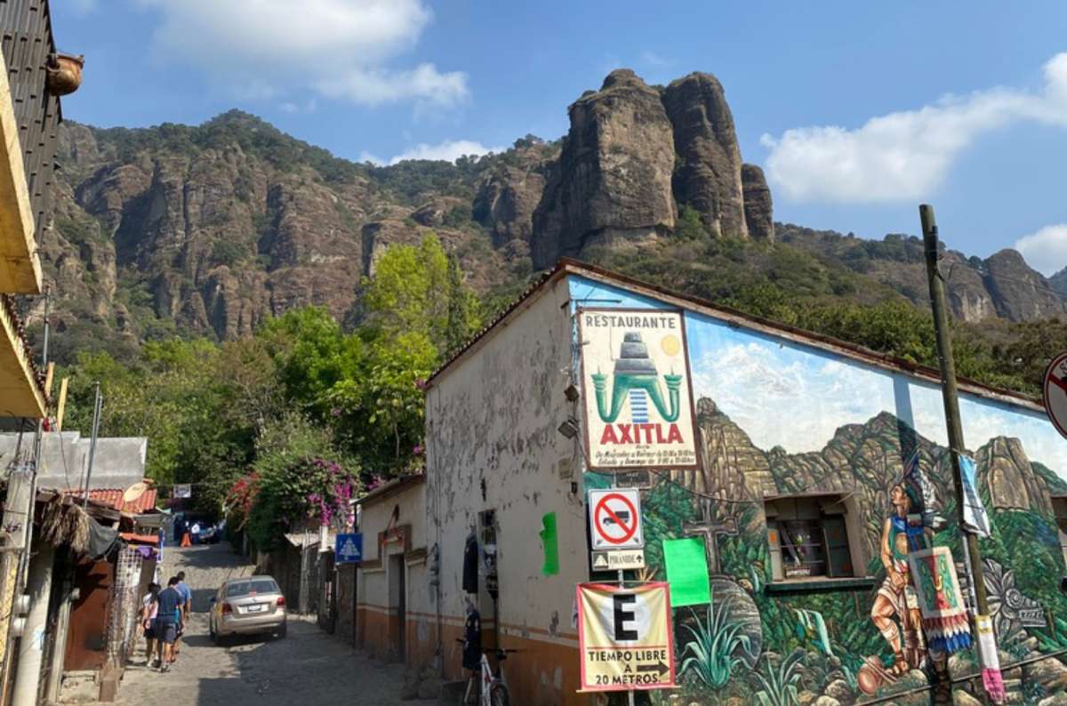 a photo of a cobblestone street with a colorful mural, a local restaurant sign, and dramatic rocky cliff formations towering above the town, Mexico, photo by Next Level of Travel