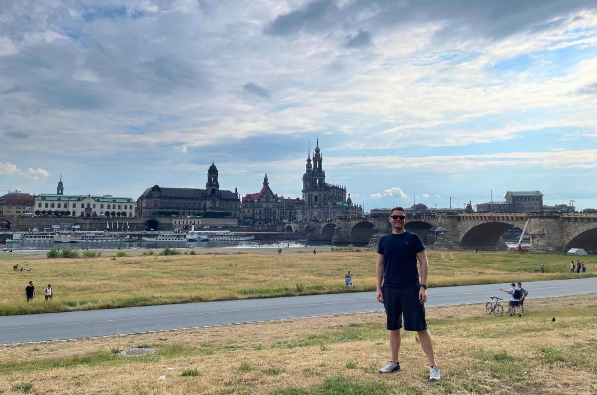 A man on a grassy riverside path in front of the Elbe River. In the background is the historic skyline of Dresden, Germany, photo by Next Level of Travel