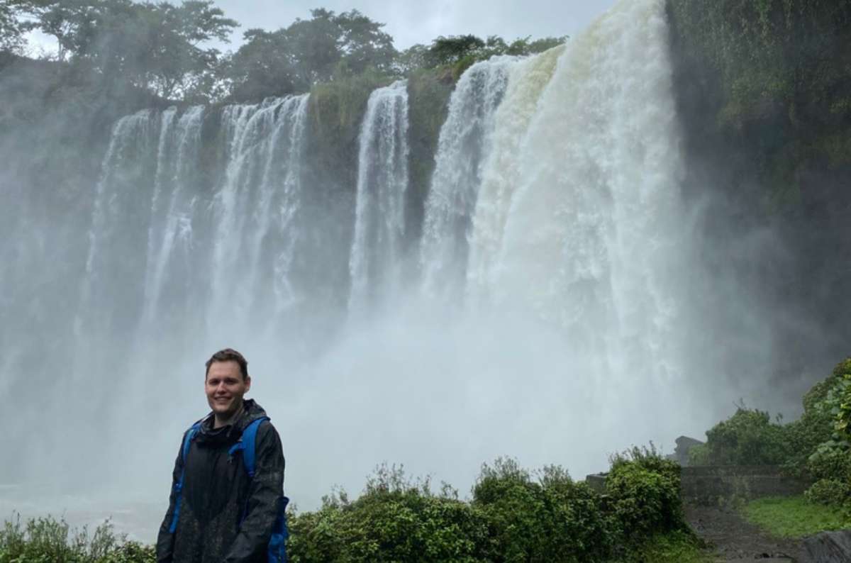 a photo of a tourist in a rain jacket standing in front of a wide, powerful waterfall, Mexico, photo by Next Level of Travel