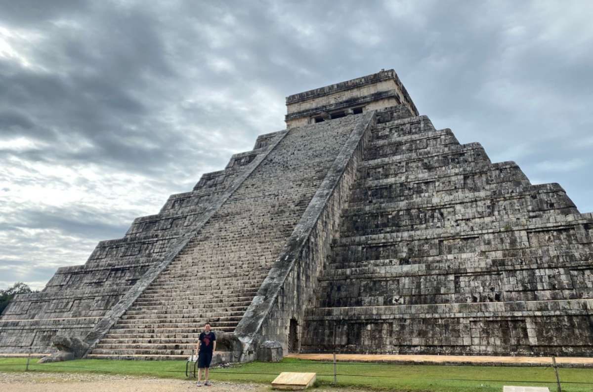 a photo of a tourist standing in front of the El Castillo pyramid at Chichén Itzá under an overcast sky, Mexico, photo by Next Level of Travel