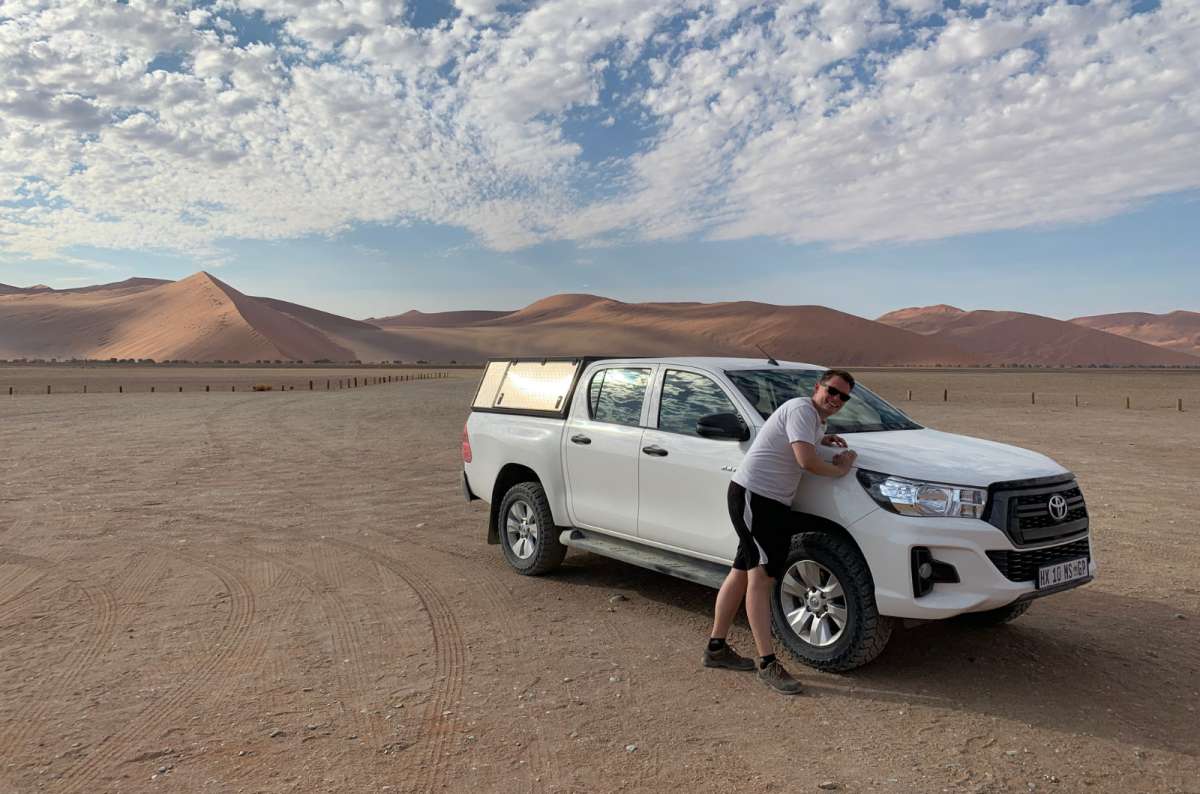 a photo of a tourist posing with a 4x4 Toyota Hilux in front of the Sossusvlei sand dunes in Namibia, photo by Next Level of Travel