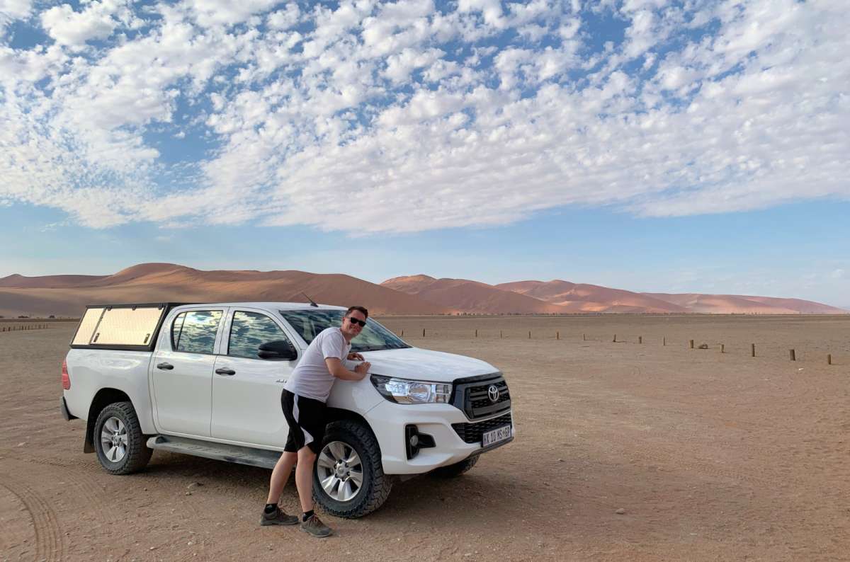 Tourist leaning on a white 4x4 pickup truck in the desert near Sossusvlei, Namib-Naukluft National Park, Namibia, photo by Next Level of Travel