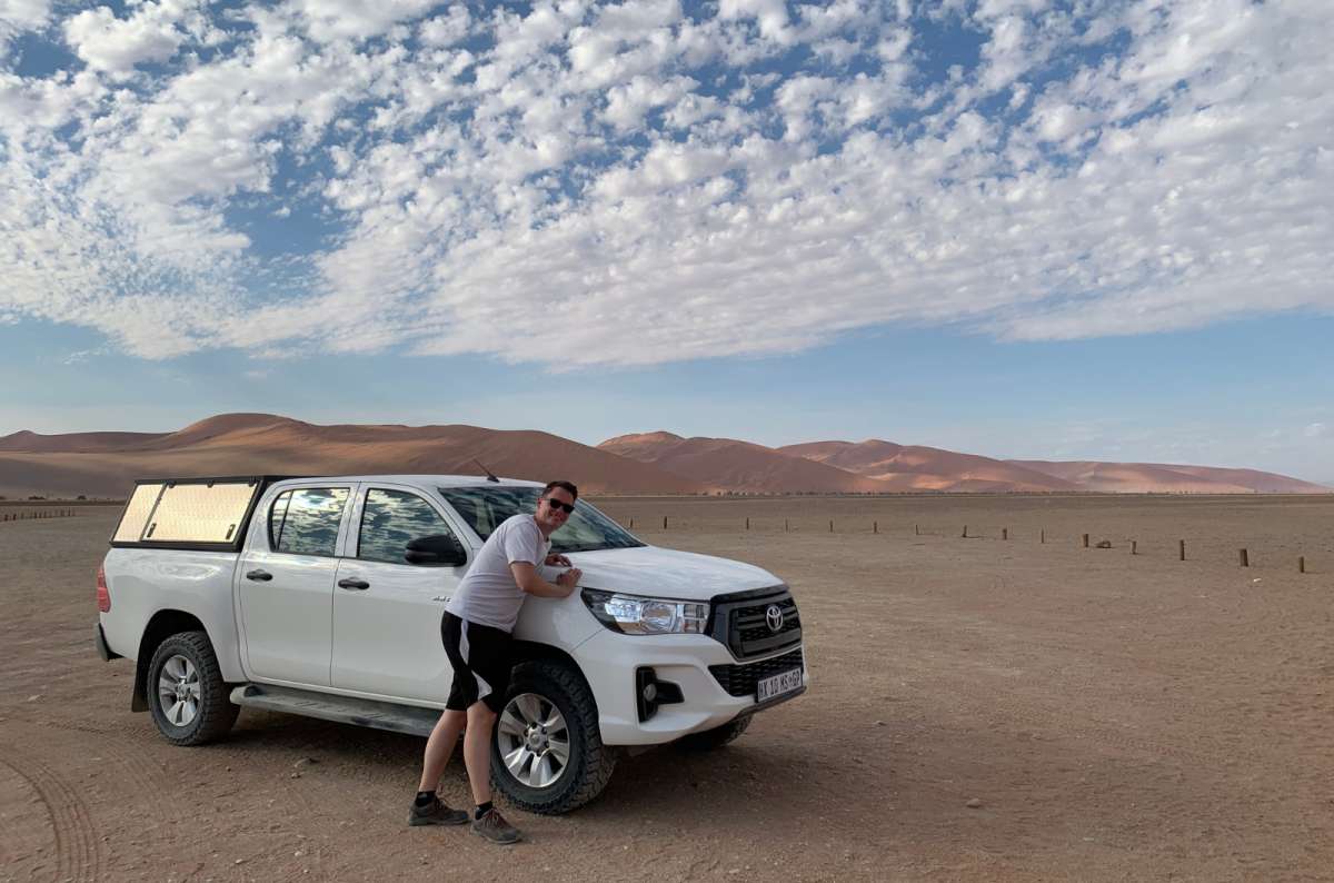 a photo of a tourist posing with a 4x4 Toyota Hilux in the Namibian desert near Sossusvlei, with large red sand dunes visible in the background, photo by Next Level of Travel