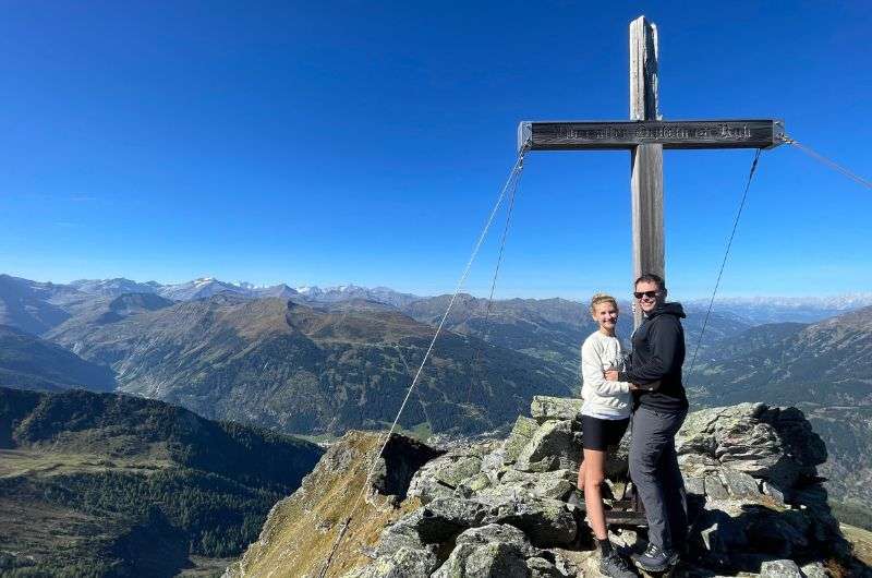 A couple standing together on the summit of Graukogel, with a wooden cross and panoramic views of the Austrian Alps, photo by Next level of travel 