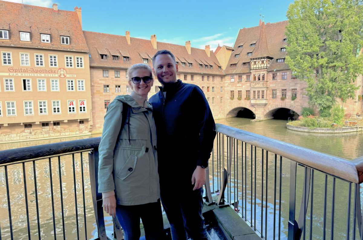 Smiling couple standing on a bridge in Nuremberg, Germany, with historic half-timbered buildings and a river in the background, photo by Next Level of Travel 