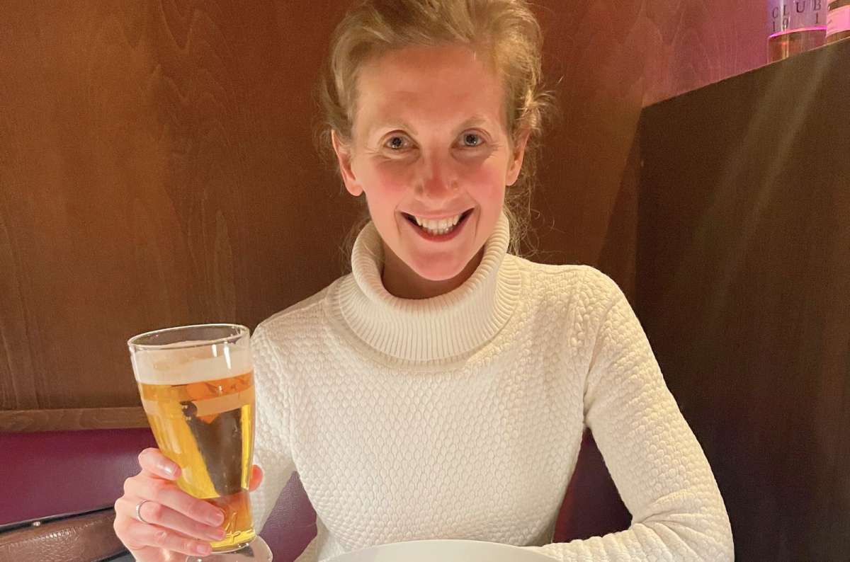 Smiling woman in a white sweater holding a glass of beer at a cozy restaurant table, Germany, photo by Next Level of Travel