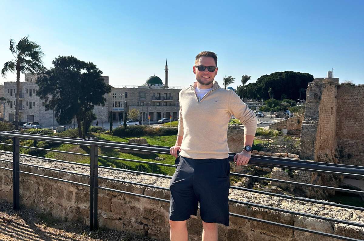 Tourist standing by the stone walls of the Citadel of Akko, Israel, with the Al-Jazzar Mosque and Old City buildings in the background, photo by Next Level of Travel