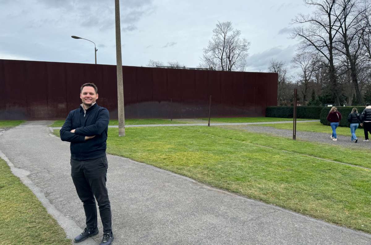 Man standing and smiling at the Berlin Wall Memorial on Bernau Straße, Germany with a preserved section of the wall and grassy no man's land in the background, photo by Next Level of Travel
