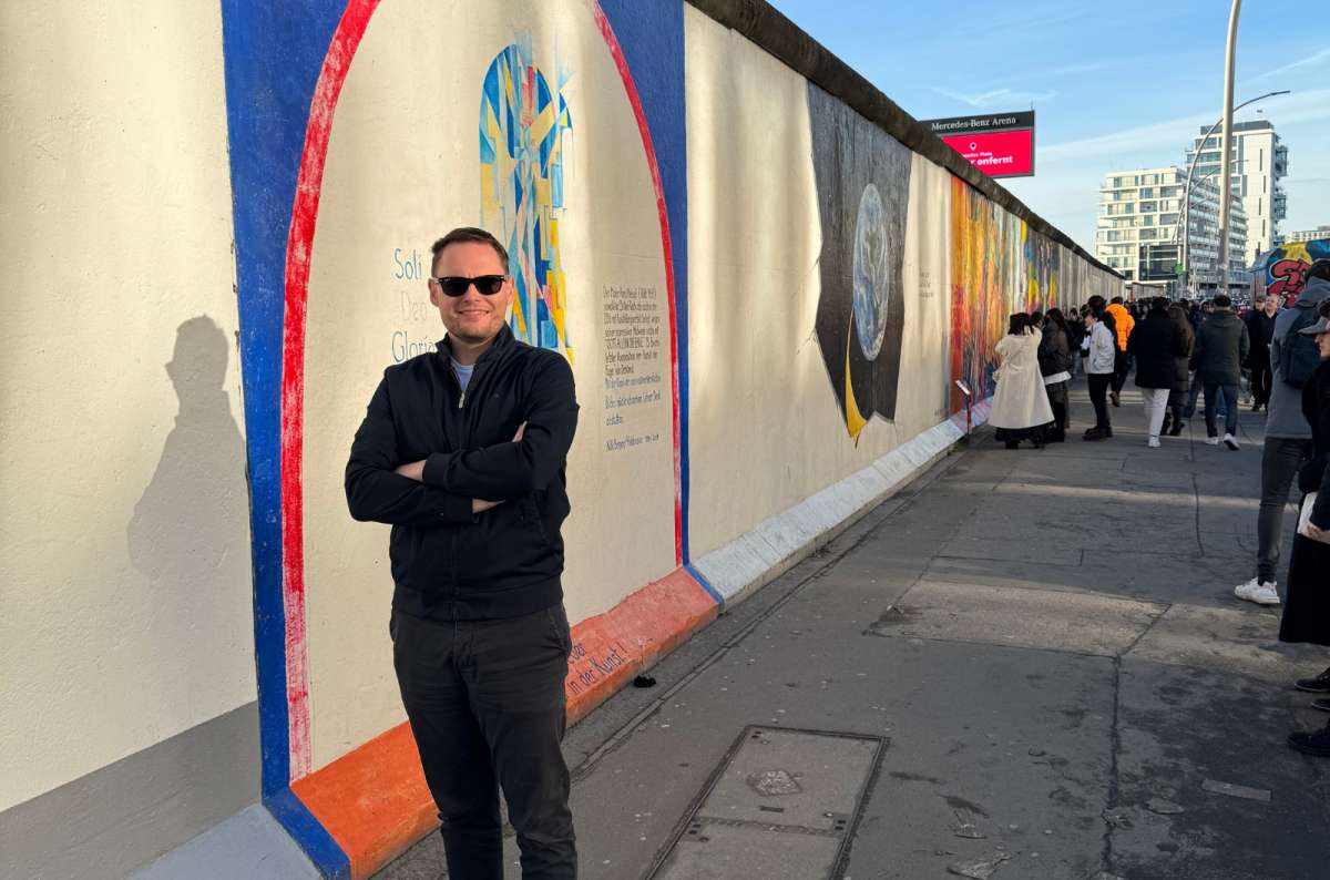 Tourist smiling in front of the East Side Gallery section of the Berlin Wall, Germany with colorful murals, photo by Next Level of Travel