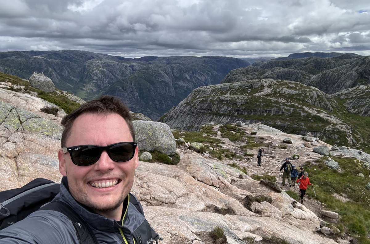 a photo of a tourist posing on the summit of a mountain hike in Lysefjord with other tourists visible in the background, Norway, photo by Next Level of Travel