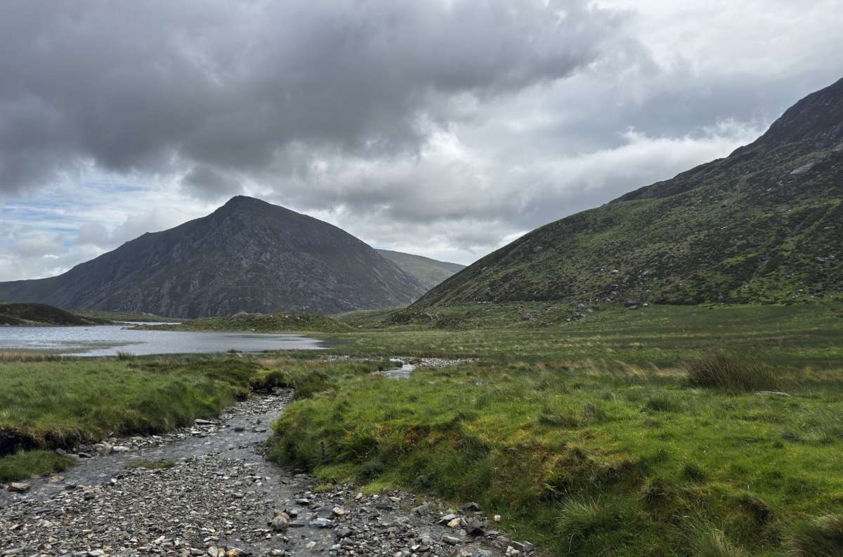 Mountain lake surrounded by green hills under dramatic clouds in Wales, a photo by Next Level of Travel