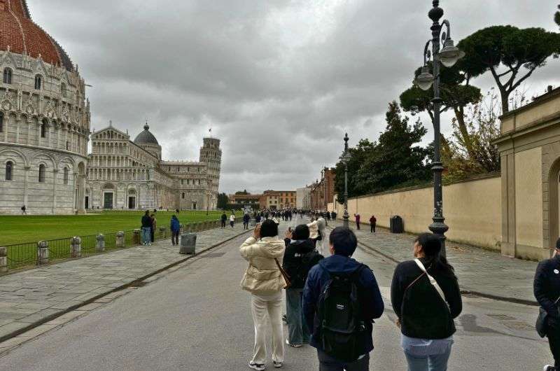 Tourists walking toward the Leaning Tower of Pisa on a cloudy day, with the Baptistery and Cathedral visible in Piazza dei Miracoli, Italy, photo by Next level of travel