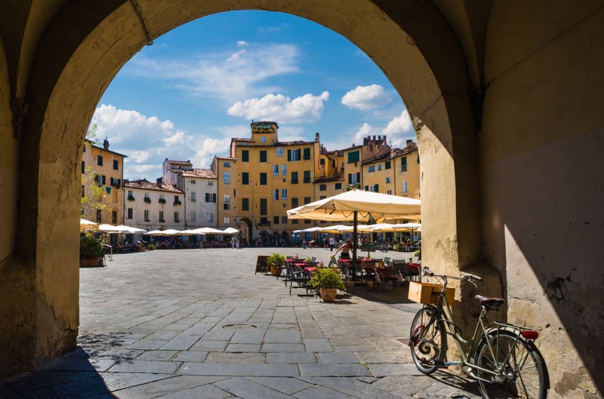 View of Lucca’s Piazza dell’Anfiteatro through a stone archway, with yellow buildings, outdoor café tables, and a bicycle leaning against the wall in the foreground