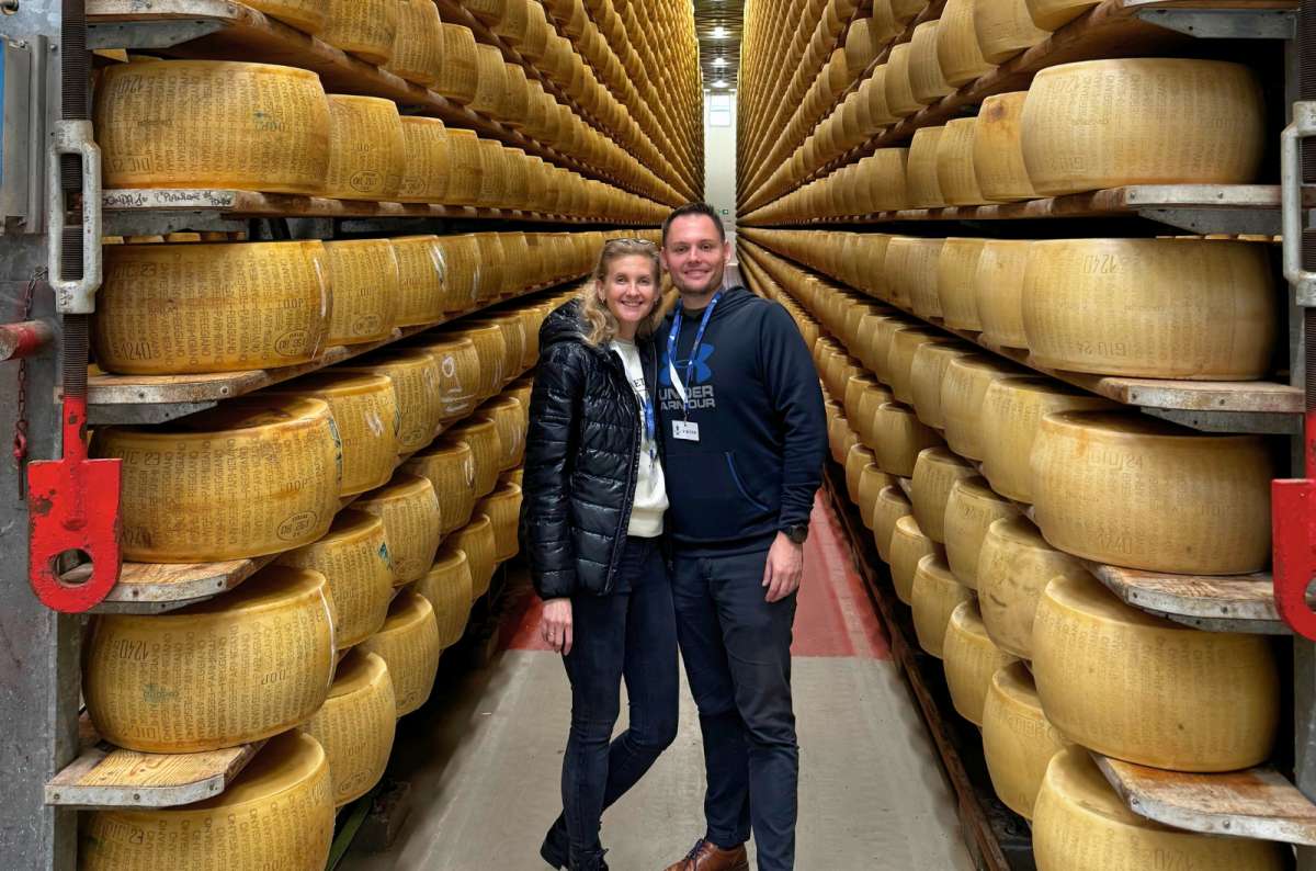 A smiling couple standing between towering rows of aging Parmigiano Reggiano wheels at the 4 Madonne Cheese Factory in Modena in Emilia Romagna, Italy, photo by Next Level of Travel