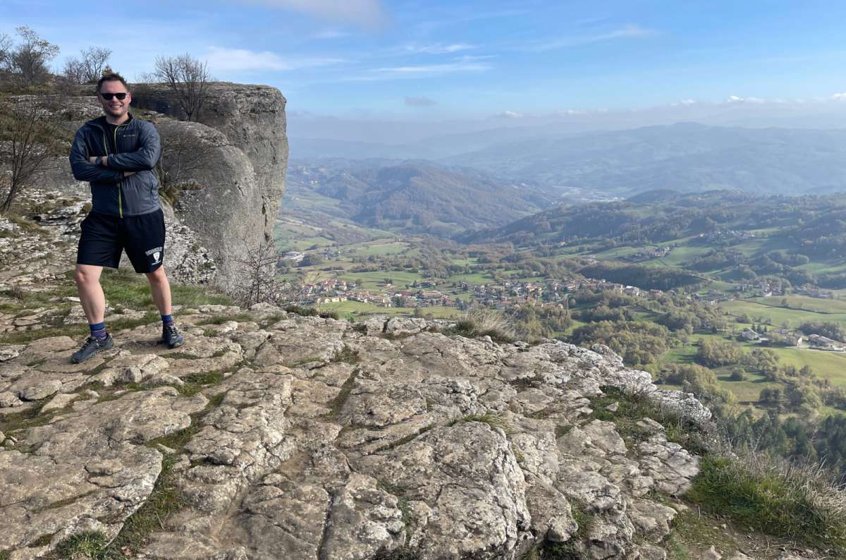Man standing on a rocky cliff at Pietra di Bismantova, Italy, overlooking rolling hills and a patchwork of fields and villages, photo by Next Level of Travel