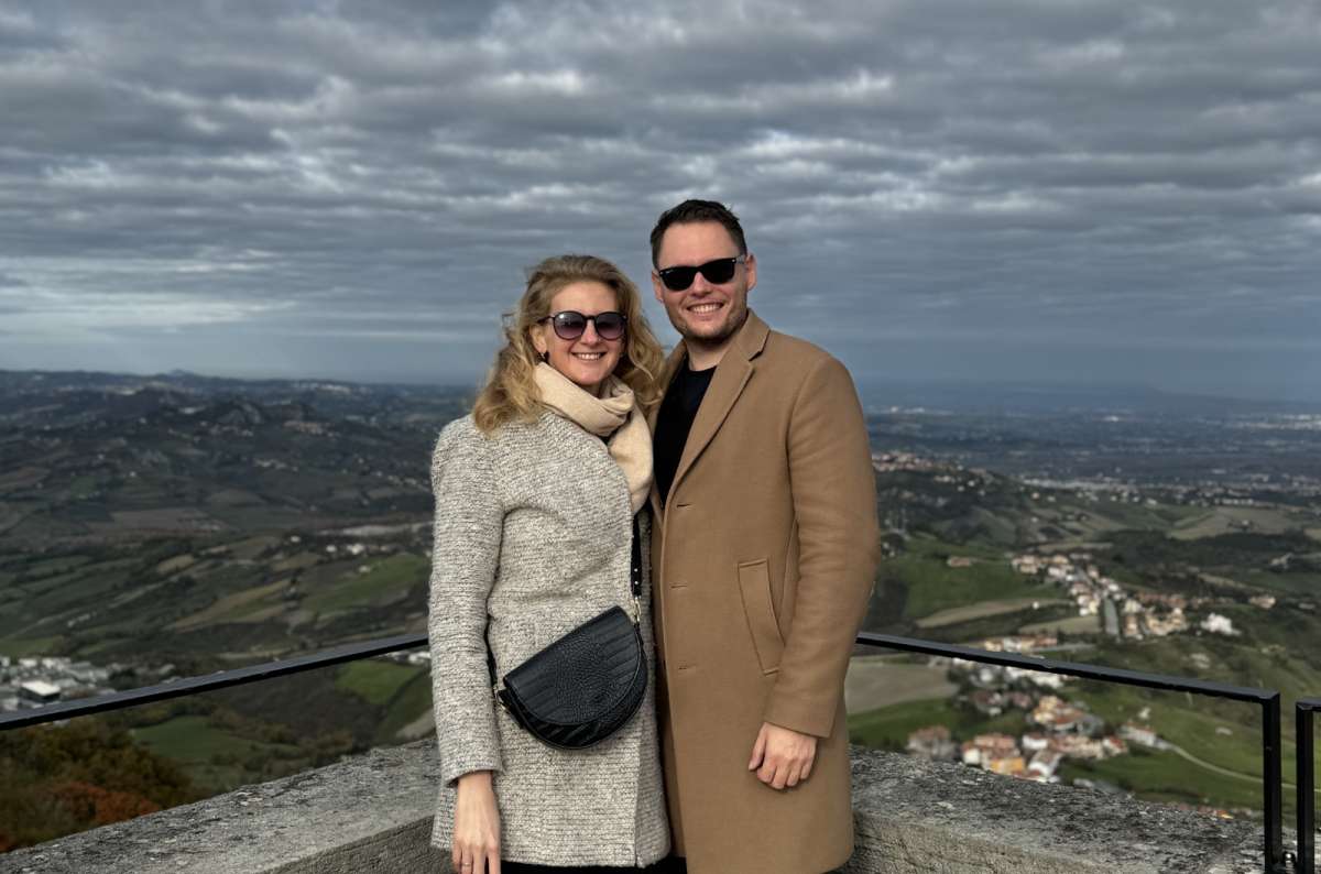 Smiling couple in coats posing at a scenic viewpoint in San Marino, Italy, with rolling green hills and scattered villages in the background, photo by Next Level of Travel