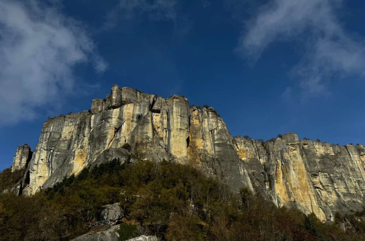 View of the dramatic limestone cliffs of Pietra di Bismantova, Italy,  rising above a forested hillside under a clear blue sky, photo by Next Level of Travel