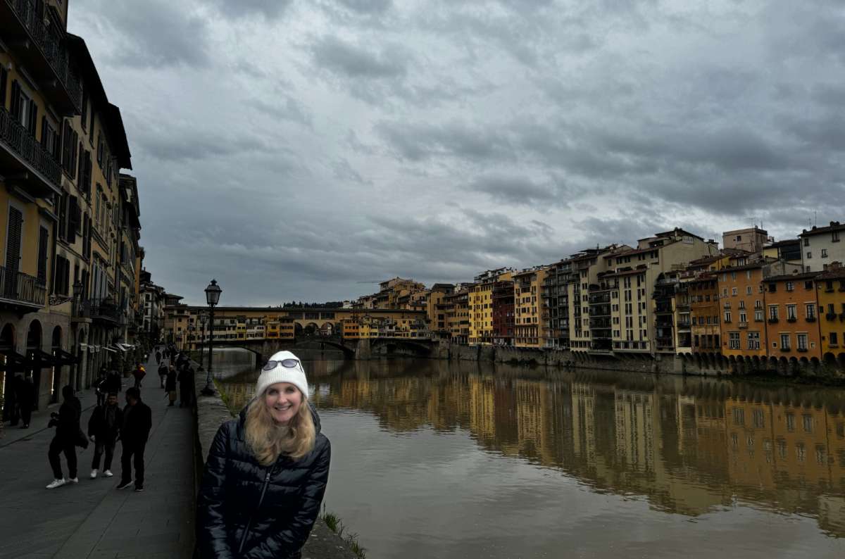 Smiling tourist standing by the Arno River with Ponte Vecchio’s colorful shops and arches in the background, Florence, Italy, photo by Next Level of Travel