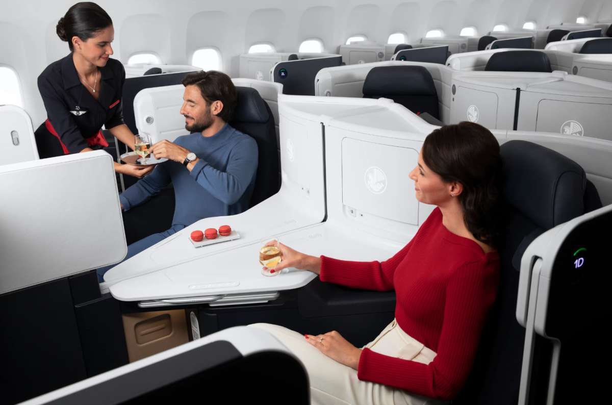 Business Class cabin with spacious seats, flight attendant serving drinks, and passengers enjoying champagne and macarons during a flight