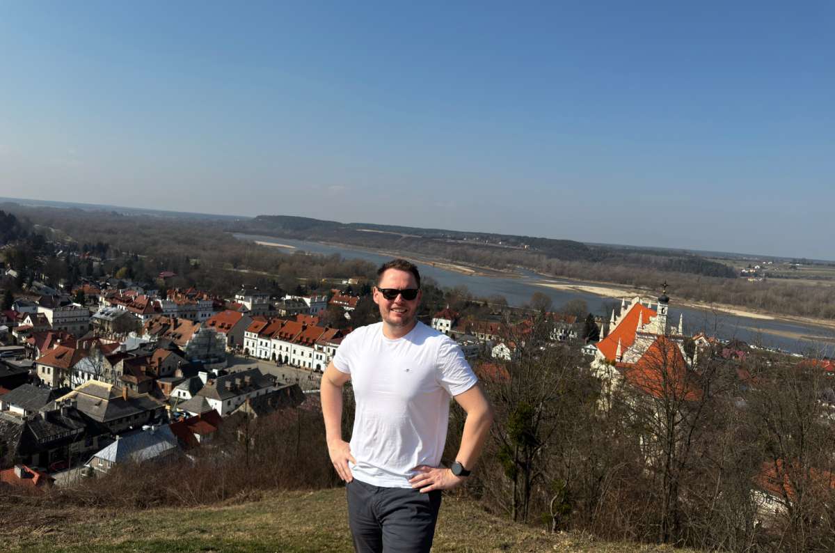 A photo of a tourist posing in on top of a hill with the village of Kazimierz Dolny visible in the background, Poland, photo by Next Level of Travels