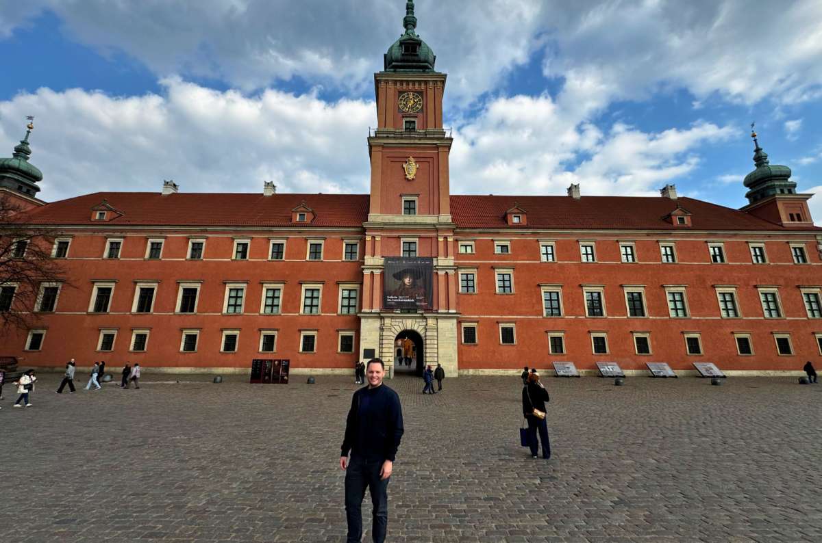 Traveler standing in front of the red-brick Royal Castle in Warsaw, Poland, on Castle Square under a partly cloudy sky, photo by Next Level of Travel