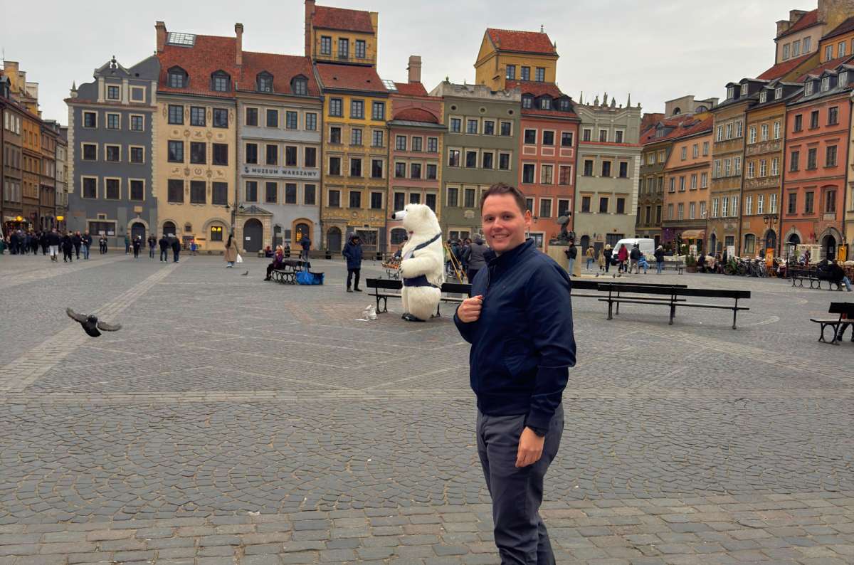 Traveler standing in Warsaw Old Town Market Square with colorful historic buildings and the Syrenka statue in the background, Poland, photo by Next Level of Travel