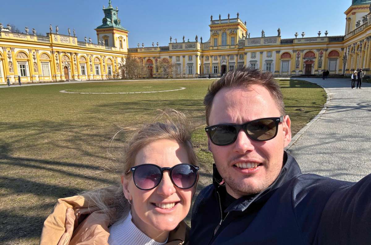 Two visitors taking a selfie in front of the yellow Baroque-style Wilanów Palace on a sunny day in Warsaw, Poland, photo by Next Level of Travel