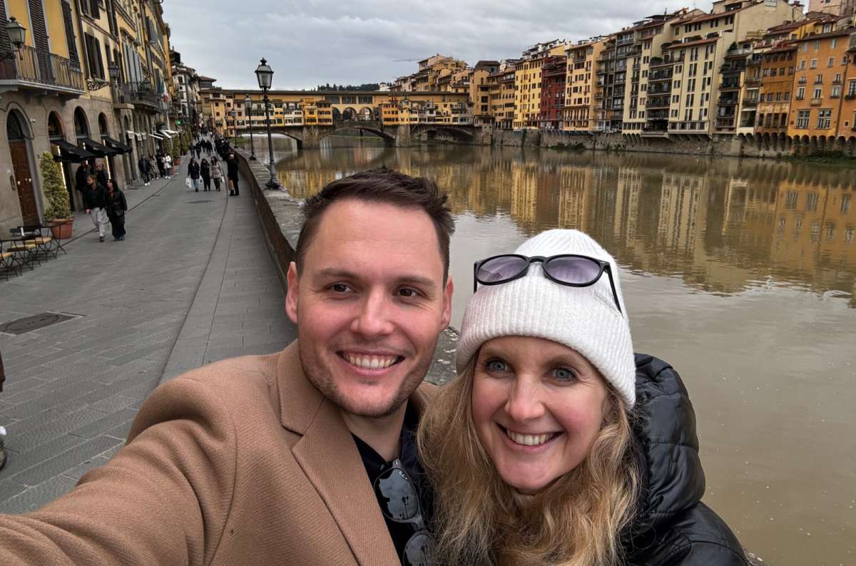 Couple taking a selfie by the Arno River in Florence, Italy, with Ponte Vecchio and colorful riverside buildings in the background, photo by Next Level of Travel