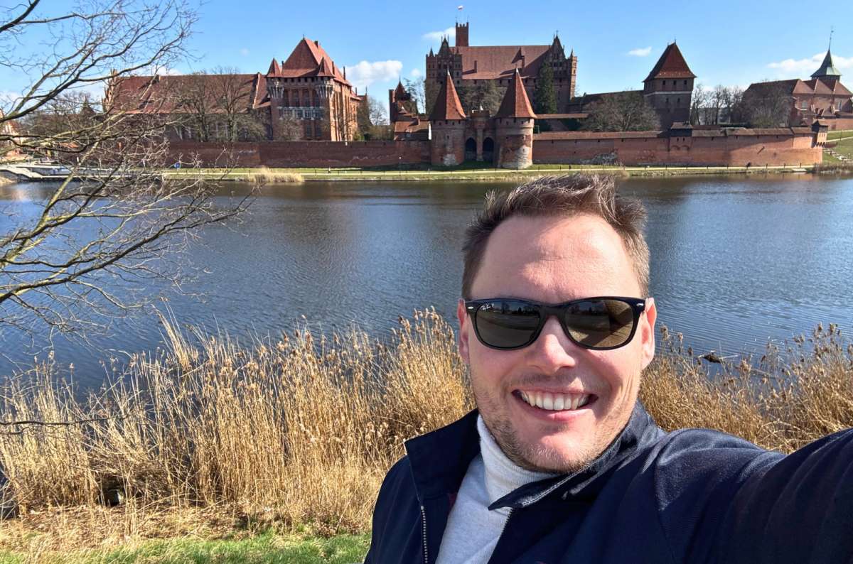 Traveler taking a selfie with Malbork Castle across the Nogat River in Malbork, Poland, a must-visit on any Poland itinerary, photo by Next Level of Travel