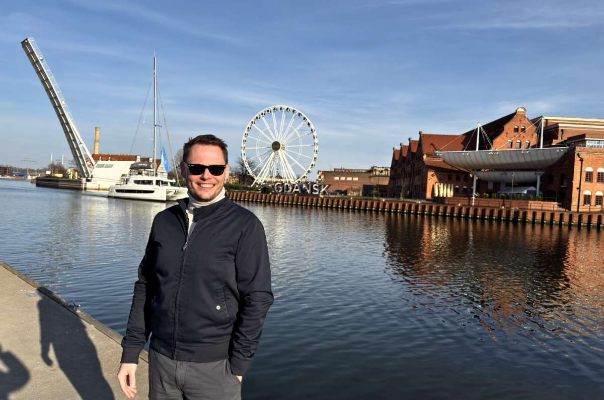 Man standing by the Motława River in Gdansk, Poland with the Gdansk sign, Ferris wheel, and waterfront buildings in the background, photo by Next Level of Travel