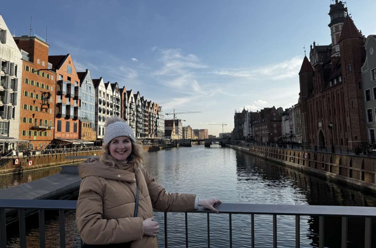 Woman standing on a bridge over the Motława River in Gdansk Old Town, Poland, with historic red-brick buildings and modern waterfront architecture, photo by Next Level of Travel