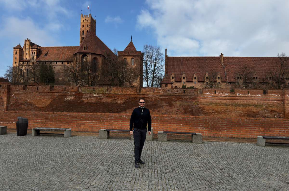 a photo of a tourist posing with the Malbork Castle in the background, photo by Next Level of Travel