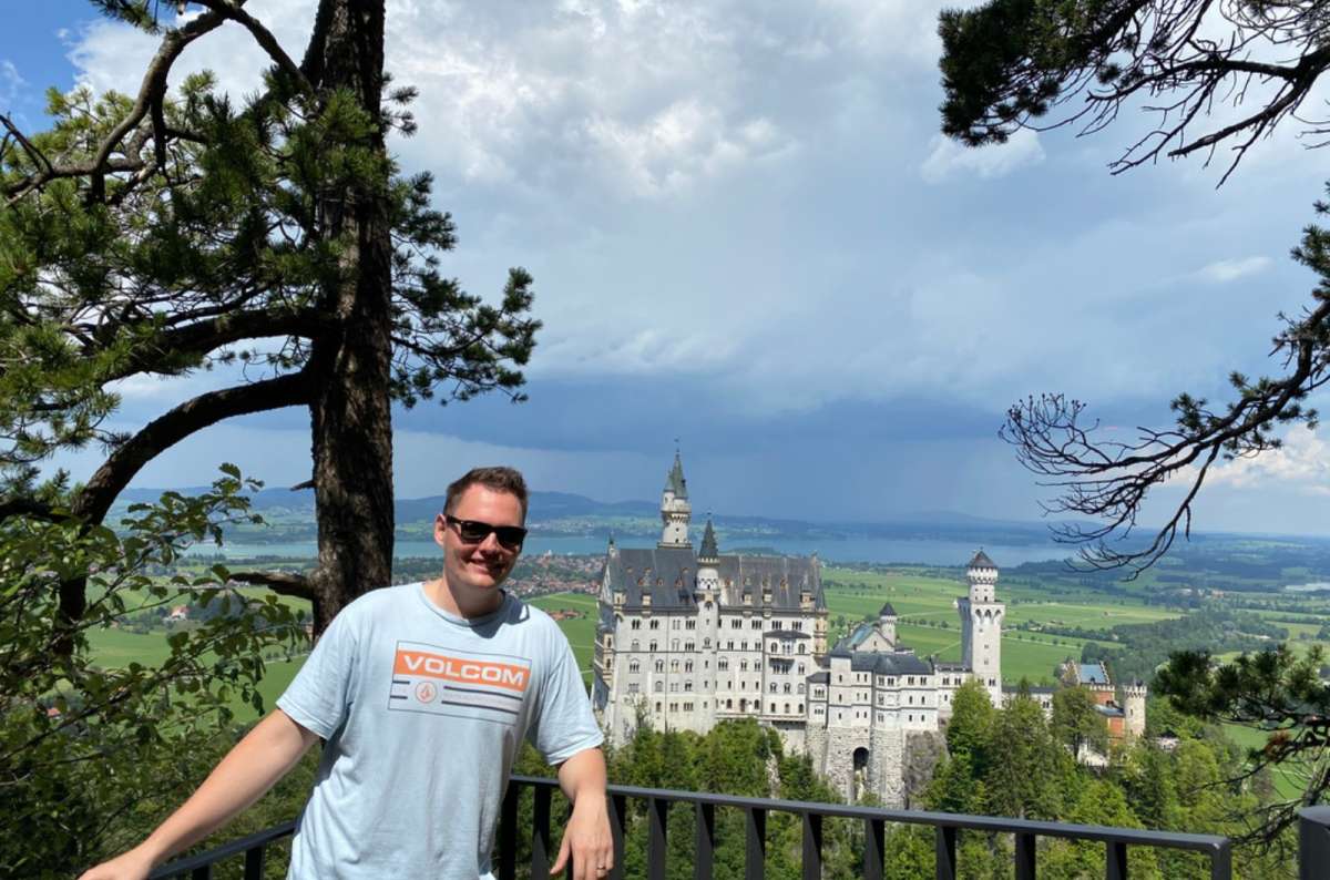 a photo of a tourist posing in front of Neuschwanstein Castle surrounded by mountains in Bavaria, Germany, photo by Next Level of Travel