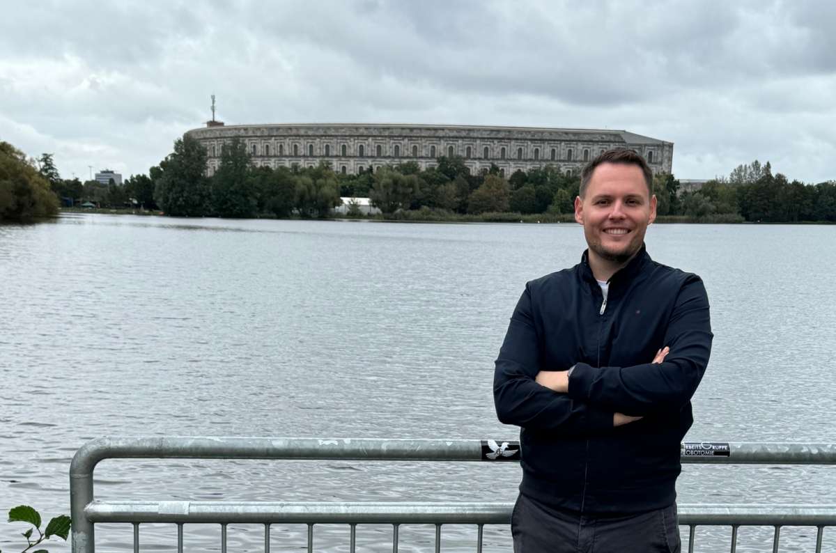 a photo of a tourist posing in front of a lake with the Nazi Rally Grounds visible in the background, Germany, photo by Next Level of Travel