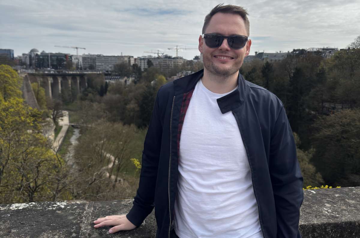 a photo of a tourist posing on the Adolphe bridge with the Valley and the modern city visible in the background, Luxembourg, photo by Next Level of Travel