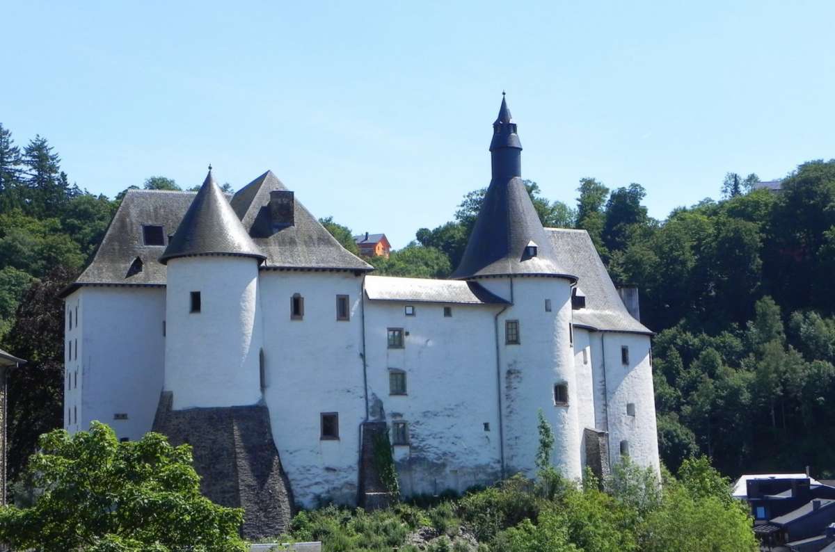 a photo of the whitewashed Clervaux Castle with its grey conical towers on a wooded hill, Clervaux, Luxembourg