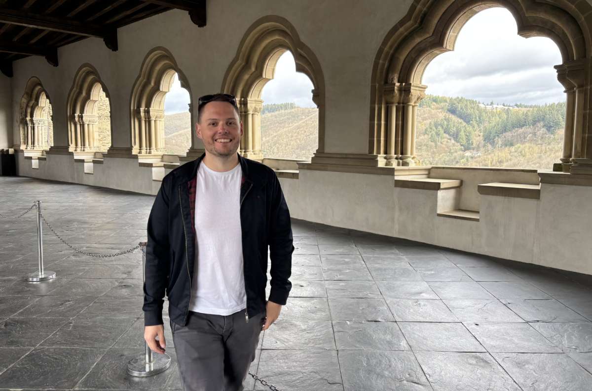 a photo of a tourist in the arcade gallery of Vianden Castle with stone arches and forested hills visible behind, Vianden Castle, Luxembourg, photo by Next Level of Travel