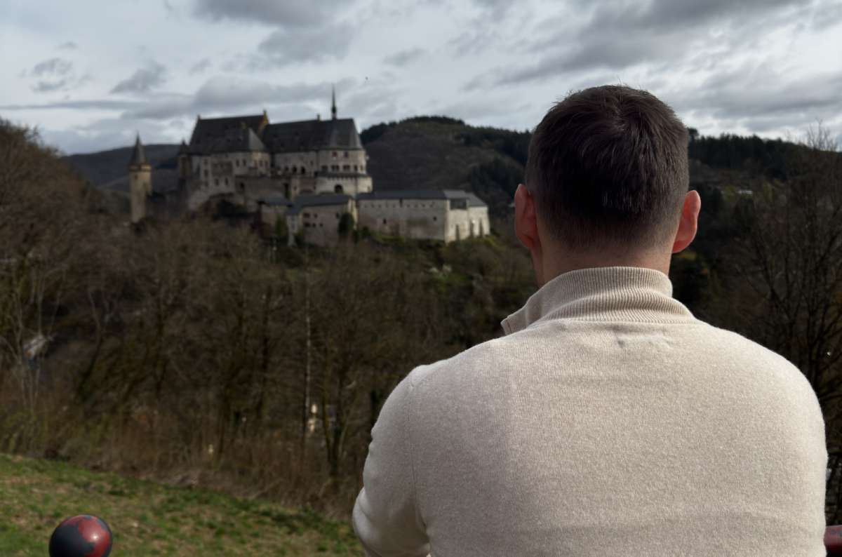 a photo of a tourist viewed from behind looking across the valley at Vianden Castle perched dramatically on the wooded hillside, Luxembourg, photo by Next Level of Travel