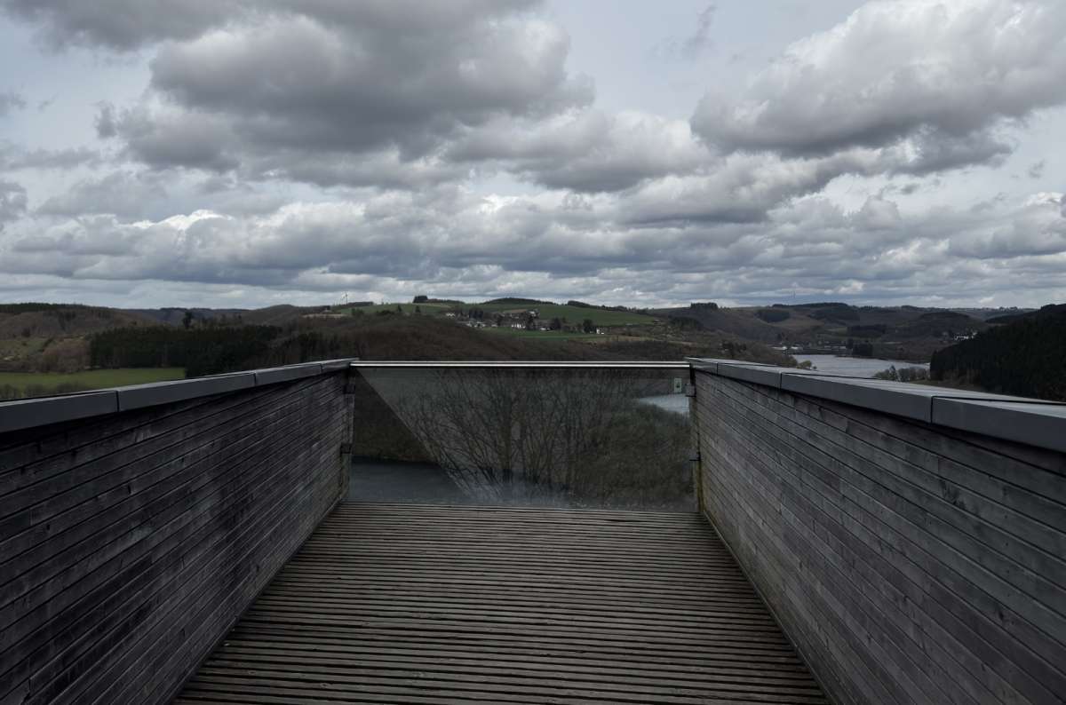 a photo of a wooden observation deck walkway overlooking the Lac de la Haute-Sure reservoir surrounded by hills and the sky, Luxembourg, photo by Next Level of Travel